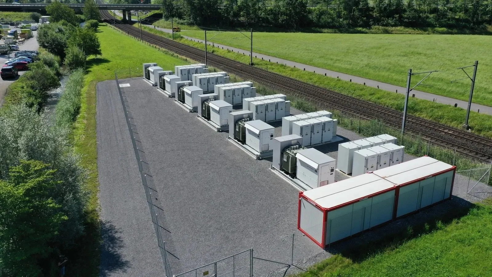 Row of containerised battery energy storage units on an outdoor pad beside a rail line